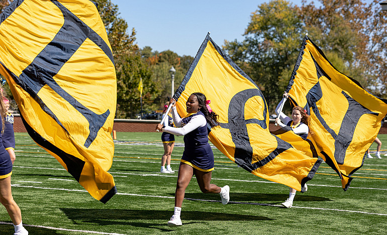 Cheerleaders carrying flags with the characters 'E,' '&' and 'H' on each